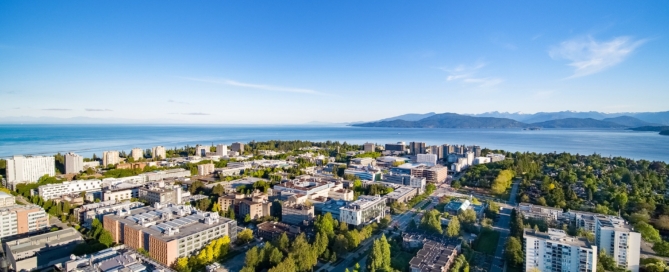 A wide cityscape under a clear blue sky with the ocean visible in the distance, capturing the vibrant environment surrounding the University of British Columbia.